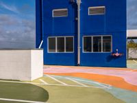 colorful painted tennis court with blue building and sky behind it in the back ground, in front of it is a parking lot