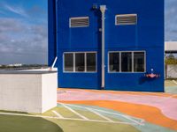 colorful painted tennis court with blue building and sky behind it in the back ground, in front of it is a parking lot