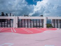 a tennis court next to an office building, under a cloudy sky with blue skies