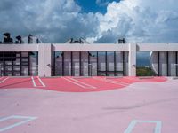 a tennis court next to an office building, under a cloudy sky with blue skies