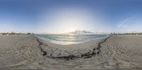 a panorama shot on the beach shows the ocean and sand around it with people, buildings and trees in the distance