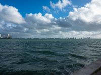 a large body of water with city buildings in the distance and clouds hovering overhead above