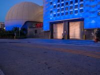 an empty road with blue lights on the building behind it in a dark night sky