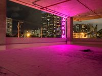 a skateboard sitting under a pink neon sign in a parking garage with tall buildings and lights on it