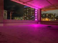 a skateboard sitting under a pink neon sign in a parking garage with tall buildings and lights on it