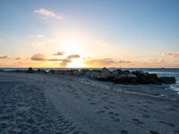 the sun rises over the ocean from an open beach with rocks and surf boards in front