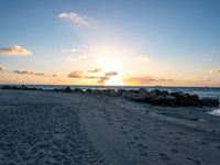 the sun rises over the ocean from an open beach with rocks and surf boards in front