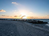 the sun rises over the ocean from an open beach with rocks and surf boards in front