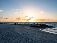 the sun rises over the ocean from an open beach with rocks and surf boards in front