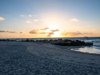 the sun rises over the ocean from an open beach with rocks and surf boards in front