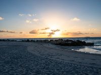 the sun rises over the ocean from an open beach with rocks and surf boards in front