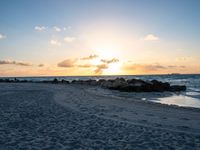 the sun rises over the ocean from an open beach with rocks and surf boards in front