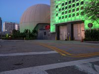 a big building with an entrance that is lighted green at night with buildings in the background