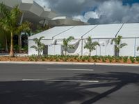 an industrial tent stands next to a road and palm trees with a gray roof and blue sky in the background