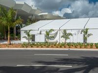 an industrial tent stands next to a road and palm trees with a gray roof and blue sky in the background