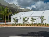 an industrial tent stands next to a road and palm trees with a gray roof and blue sky in the background