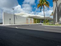 a road near a building with palm trees and clouds in the background is a white building that has a curved roof that wraps across from a small courtyard