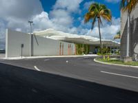 a road near a building with palm trees and clouds in the background is a white building that has a curved roof that wraps across from a small courtyard