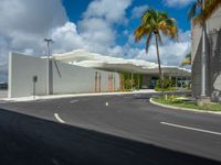 a road near a building with palm trees and clouds in the background is a white building that has a curved roof that wraps across from a small courtyard