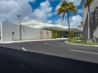 a road near a building with palm trees and clouds in the background is a white building that has a curved roof that wraps across from a small courtyard