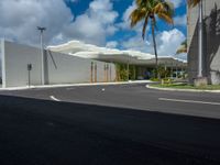 a road near a building with palm trees and clouds in the background is a white building that has a curved roof that wraps across from a small courtyard