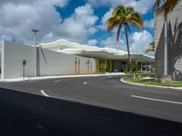 a road near a building with palm trees and clouds in the background is a white building that has a curved roof that wraps across from a small courtyard