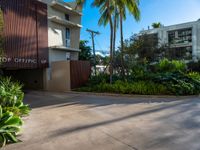 the entrance to an apartment with palms trees and bushes in the foreground, next to a concrete driveway