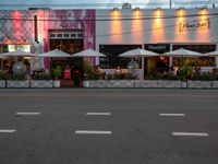 an empty street with white umbrellas and pink building with people sitting at tables and tables outside