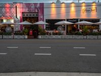 an empty street with white umbrellas and pink building with people sitting at tables and tables outside