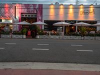 an empty street with white umbrellas and pink building with people sitting at tables and tables outside