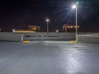 an empty parking lot with lights above it at night time on the other side of a city street