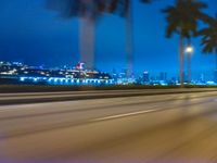 cars driving at night with tall palm trees in the foreground and skyline beyond and on the road