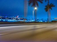 cars driving at night with tall palm trees in the foreground and skyline beyond and on the road