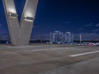 some cars parked on the asphalt at night with buildings in the background near a bridge