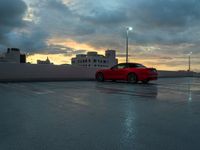 a red car parked in an empty parking lot near a cityscape at dusk