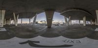 a fish eye lens showing an empty parking garage under some clouds at sunset, with buildings, and the sky visible through the gap