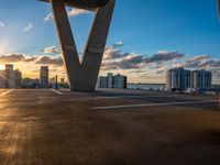 the sunset shines behind a large bridge as a car drives underneath it on a road beside large city buildings