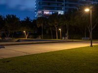 two benches sitting on the grass at night, next to an empty parking lot and tall buildings