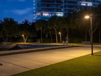 two benches sitting on the grass at night, next to an empty parking lot and tall buildings