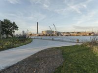 a park with some green bushes and plants next to the water and construction cranes in the background
