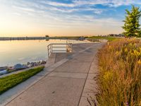 view of a wide river at sunset with grassy shoreline and pier on shore and benches to the end of the pathway