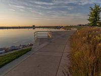 view of a wide river at sunset with grassy shoreline and pier on shore and benches to the end of the pathway