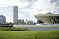 the white curved structure of an unusual building near a field of green grass, with skyscrapers in the background