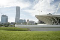 the white curved structure of an unusual building near a field of green grass, with skyscrapers in the background