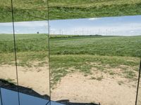 a wide mirror reflection is on the ground beside some grass and a field with wind mills