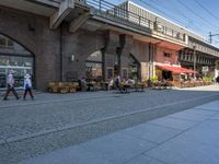 a couple of people sitting and walking on a sidewalk next to buildings near a train station