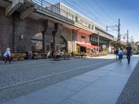 a couple of people sitting and walking on a sidewalk next to buildings near a train station