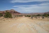 a dirt road going through the desert area with a mountainside in the distance with blue sky and clouds in the distance