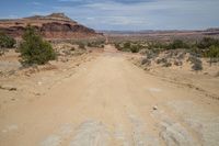 a dirt road going through the desert area with a mountainside in the distance with blue sky and clouds in the distance