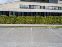 there is a parking lot with a large building and hedges in the background next to a street